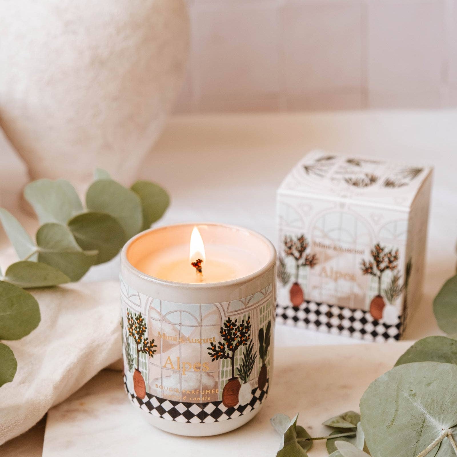 Mimi and August Candle with nature-themed design on a surface with eucalyptus leaves