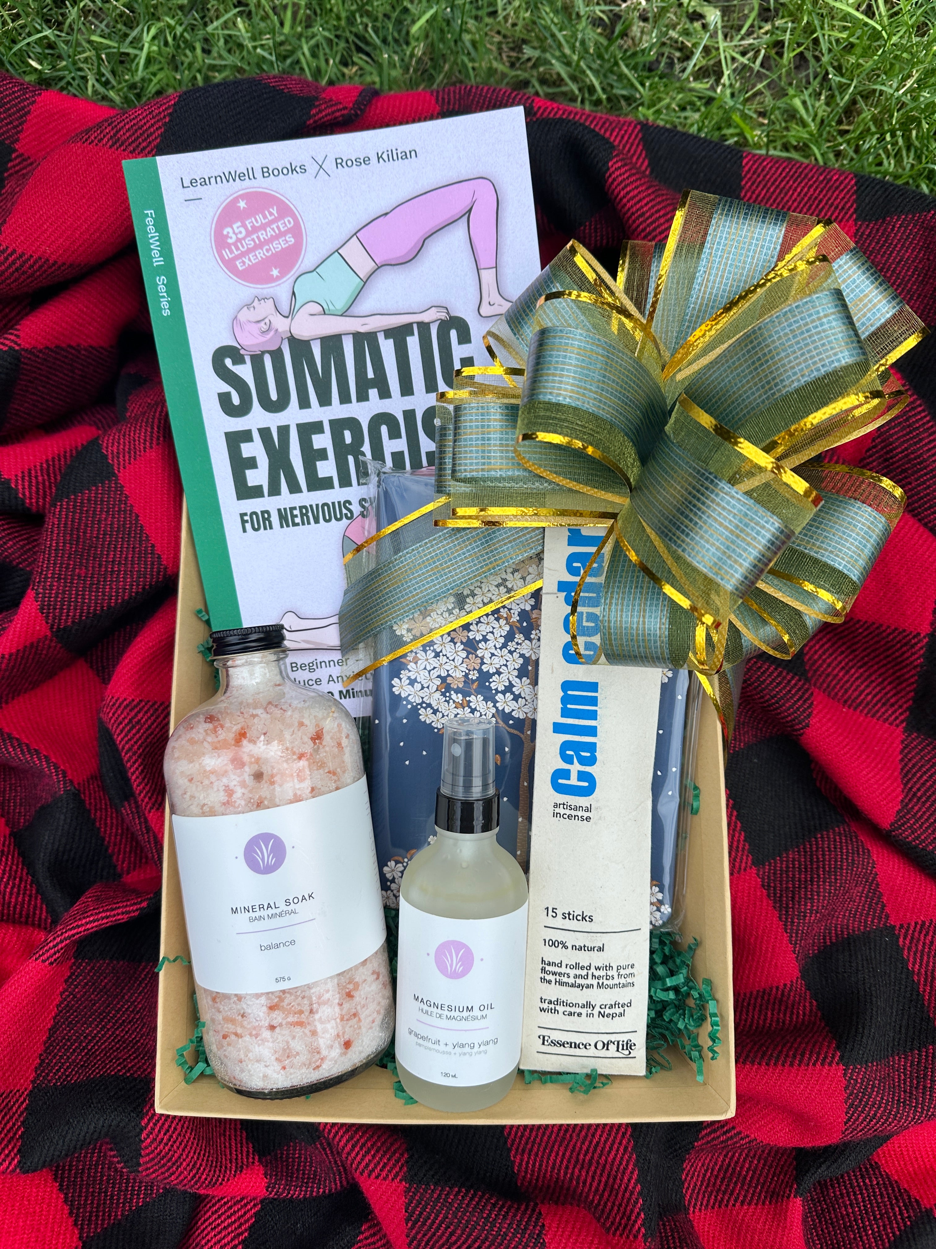 Green Beauty Curator Gift box with bath products and a book on a red and black checkered blanket outdoors.