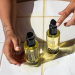 Two bottles of Om Organics oil on a tiled surface with hands interacting with them.