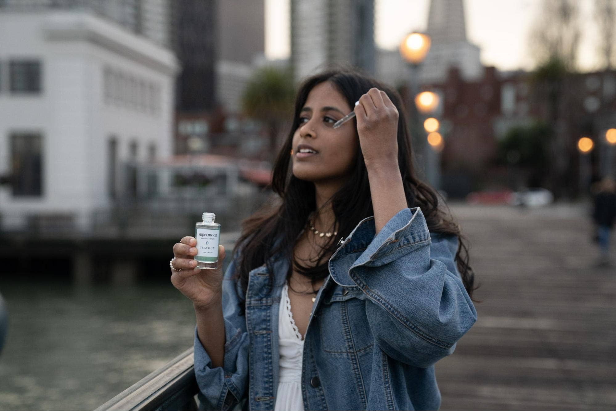 Woman holding a bottle of Graydon Supermoon serum outdoors with blurred cityscape in the background