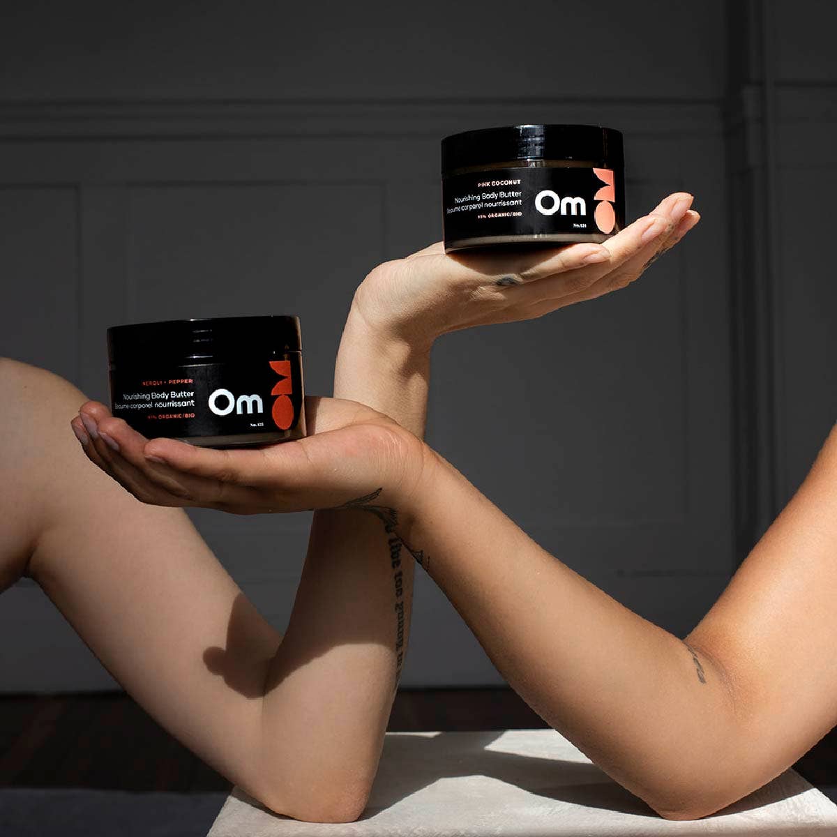 Two hands holding containers of Om body butter against a dark background
