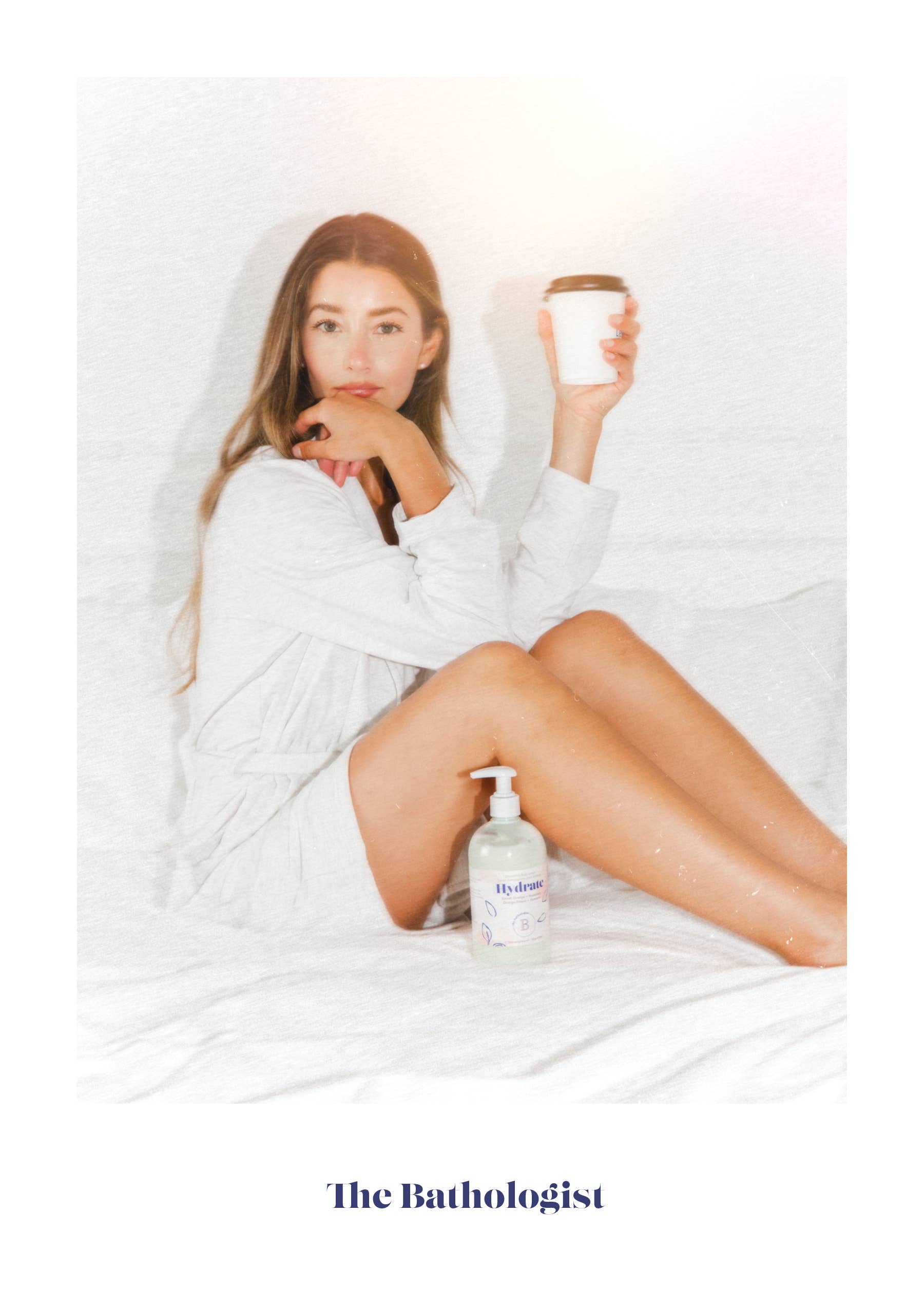 Woman in a white robe holding a cup with lotion 'The Bathologist' branding.