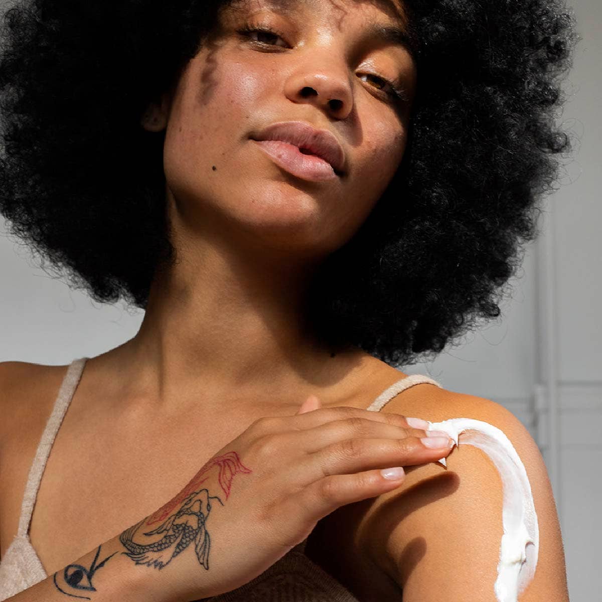 Woman with tattoos on her arm, wearing a white tank top applying Om Organics body butter against a neutral background.