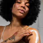 Woman with tattoos on her arm, wearing a white tank top applying Om Organics body butter against a neutral background.