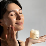 Woman applying Om Organics cream to her face with a jar of cream in her hand, against a neutral background.