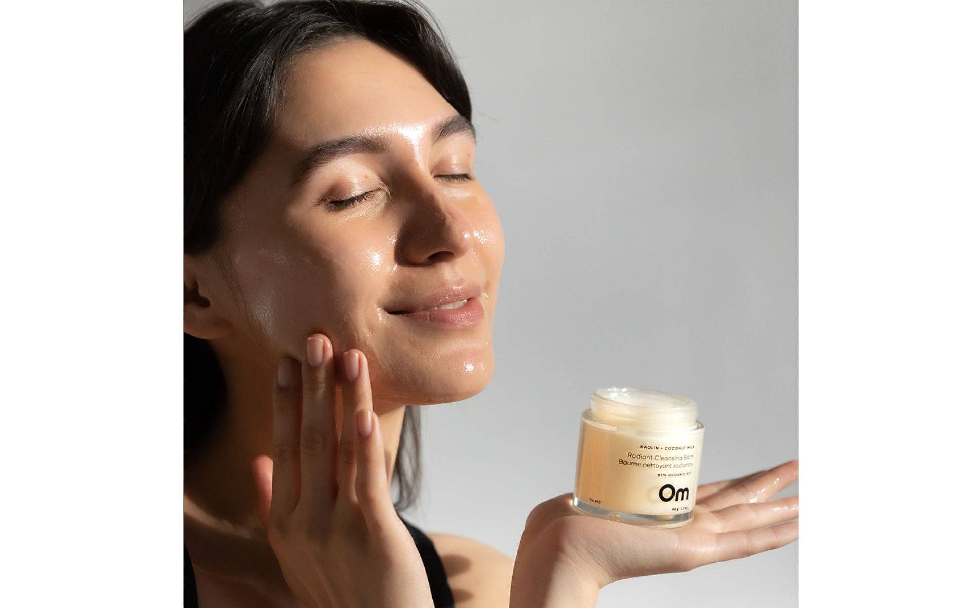 Woman applying Om Organics cream to her face with a jar of cream in her hand, against a neutral background.