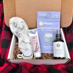 Green Beauty Curator Box of self-care items including a silk face mask, a bottle of magnesium spray, and a box of 'Superfood Latte' on a red and black checkered blanket.