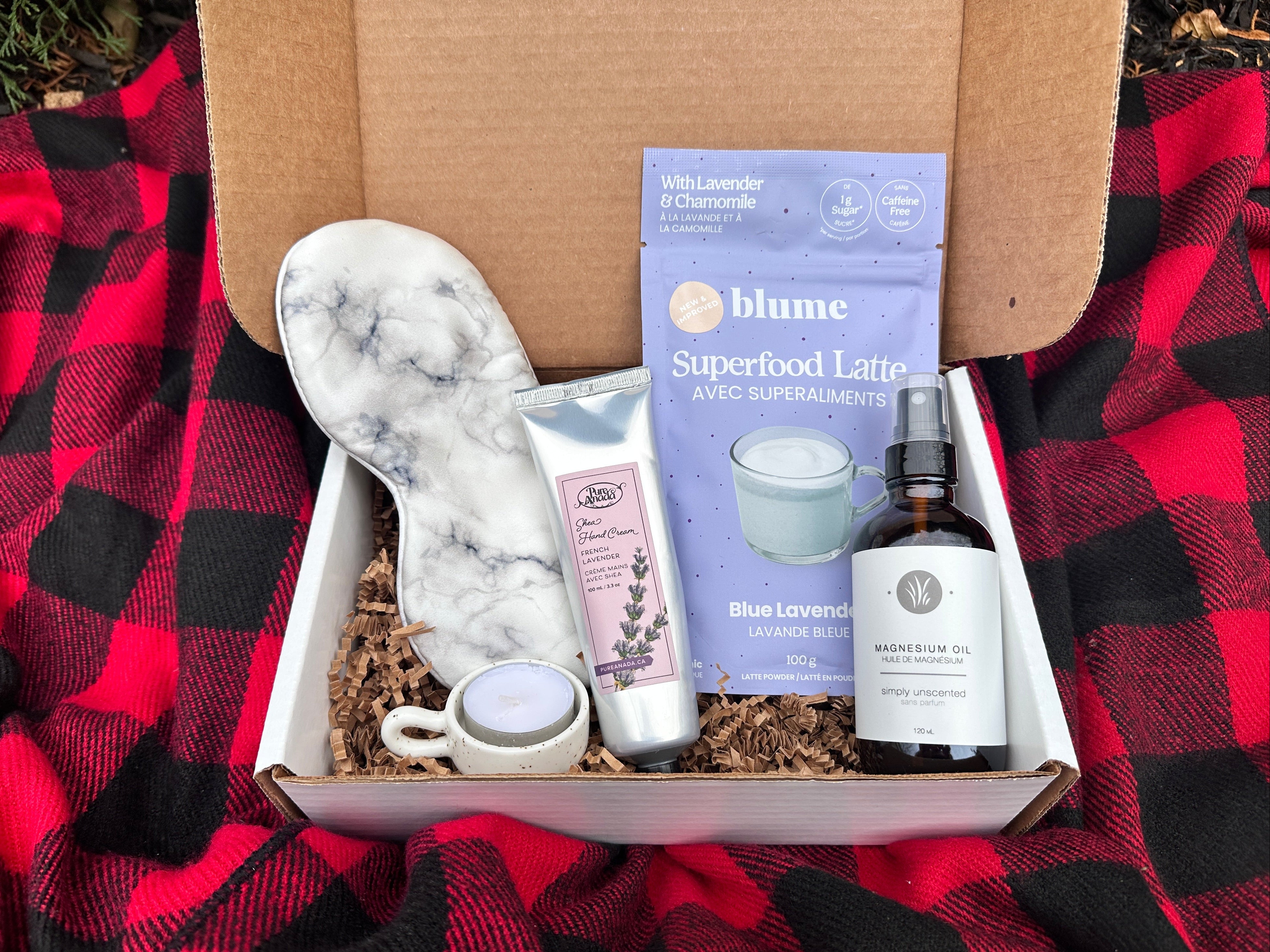 Green Beauty Curator Box of self-care items including a silk face mask, a bottle of magnesium spray, and a box of 'Superfood Latte' on a red and black checkered blanket.