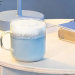 Clear mug with blue liquid and foam on a light surface with a book in the background