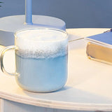 Clear mug with blue liquid and foam on a light surface with a book in the background