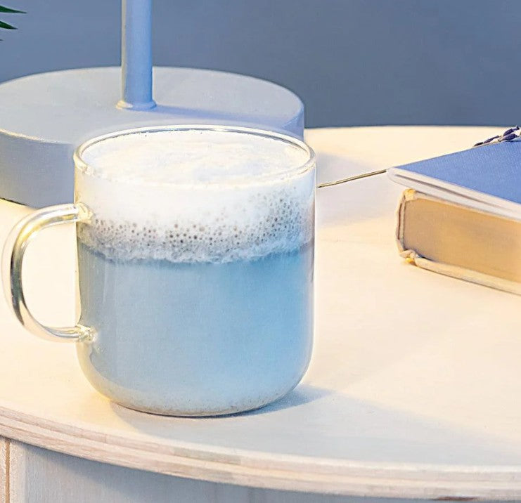 Clear mug with blue liquid and foam on a light surface with a book in the background