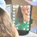 Person applying makeup in front of a mirror with lights, wearing a green sweater.