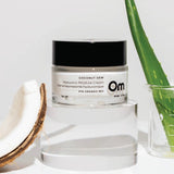 Om Organics Coconut Dew cream with a coconut and aloe vera leaf on a light background. 
