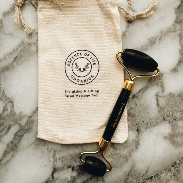 Facial roller with black stones on a marble surface next to a Essence of Life Organics branded pouch.