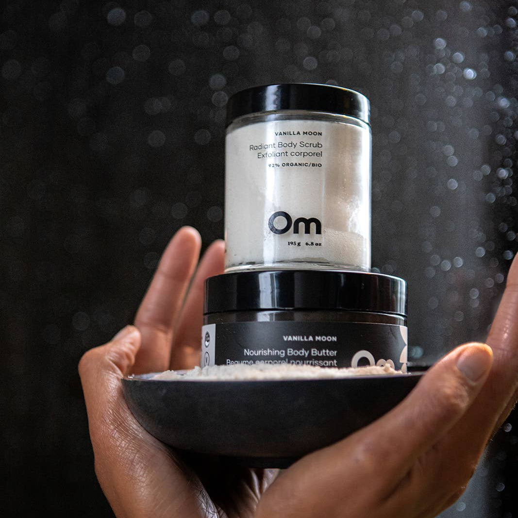 Two jars of Om Organics vanilla moon body butter held by hands against a dark background