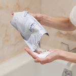 Person holding a package of The Bathologist bath salts with a soapy hand in a bathroom setting