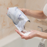Person holding a package of The Bathologist bath salts with a soapy hand in a bathroom setting