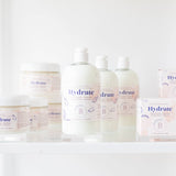 The Bathologist Hydrate skincare products on a white surface with a plant in the foreground