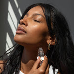 Woman applying Om perfume roll on to her neck with a bottle against a neutral background